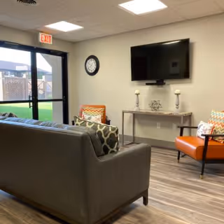A cozy living room area with a gray sofa and two orange armchairs with patterned cushions. A flat-screen TV is mounted on the wall above a console table decorated with two candlesticks and a decorative item. A clock is mounted on the wall near a glass door that leads outside to a fenced yard.