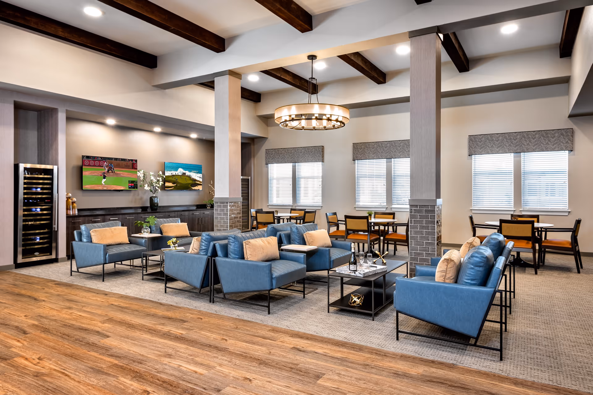 A spacious and modern common area in a senior living facility featuring blue armchairs with beige cushions arranged around black coffee tables. The room has wooden ceiling beams, a large circular chandelier, and multiple windows with blinds. In the background, there are dining tables with chairs and two flat-screen TVs mounted on the wall above a long cabinet. The floor transitions from wood to carpet in the seating area.