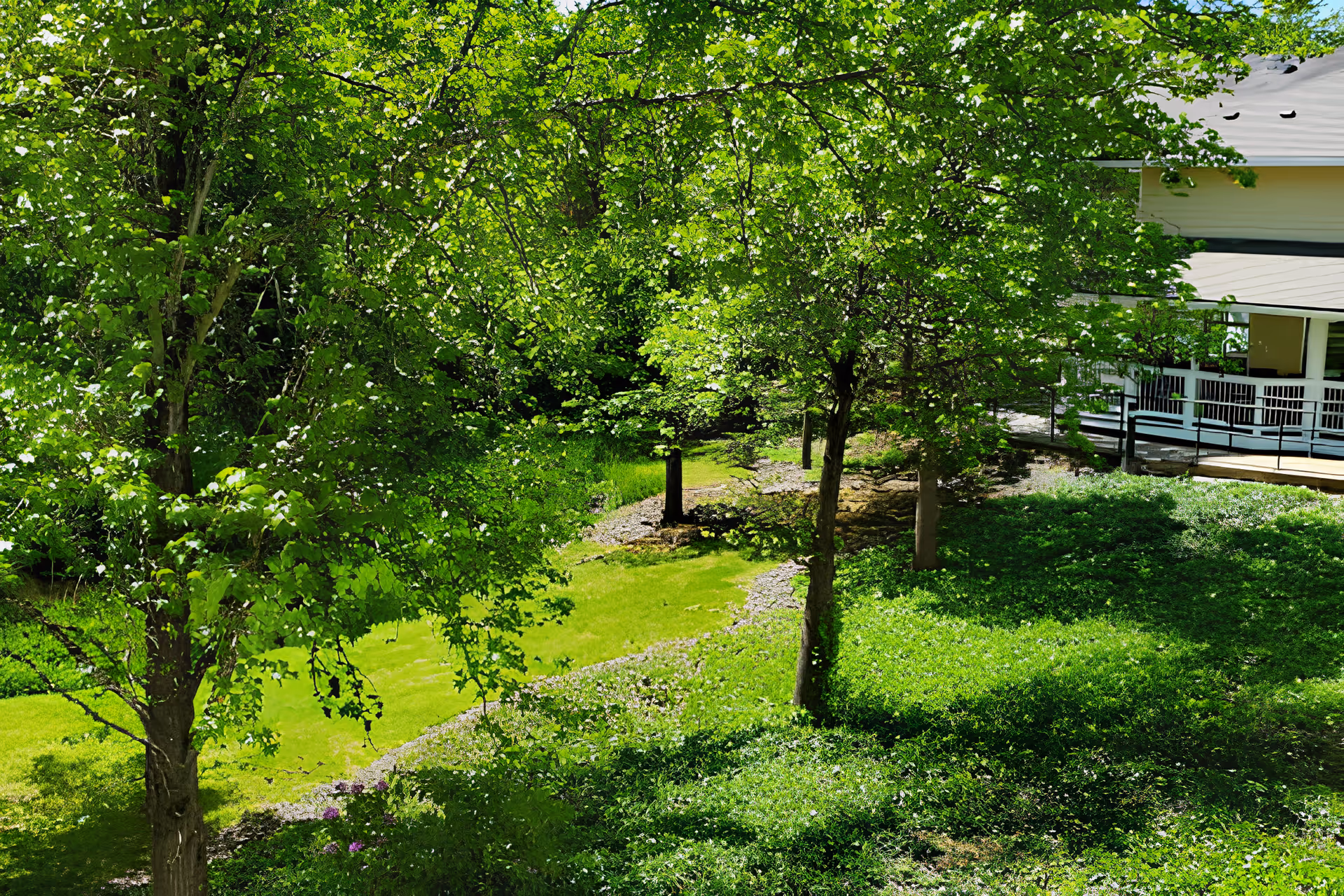 A lush green garden area with several trees and a well-maintained grassy lawn. A building with a covered porch and white railings is partially visible on the right side of the image.