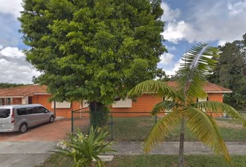 Single-story orange building with a red-tiled roof, partially obscured by a large leafy tree and a palm tree in the front yard. A silver van is parked on the driveway to the left. The sky is partly cloudy.