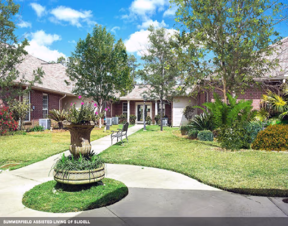 Outdoor courtyard area of Summerfield Senior Living featuring a circular planter with flowers in the center, surrounded by a concrete walkway, green grass, trees, and benches. Brick buildings with windows and a door are visible in the background under a blue sky with some clouds.