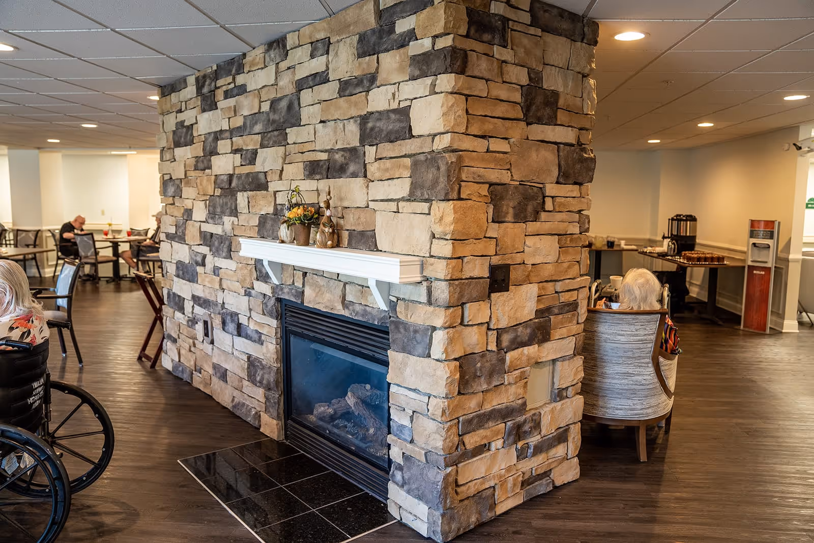 Interior view of a senior living facility common area with a stone fireplace in the center. Several elderly residents are seated around the room, some in wheelchairs and others in chairs. The room has wooden flooring and a cozy atmosphere with soft lighting.