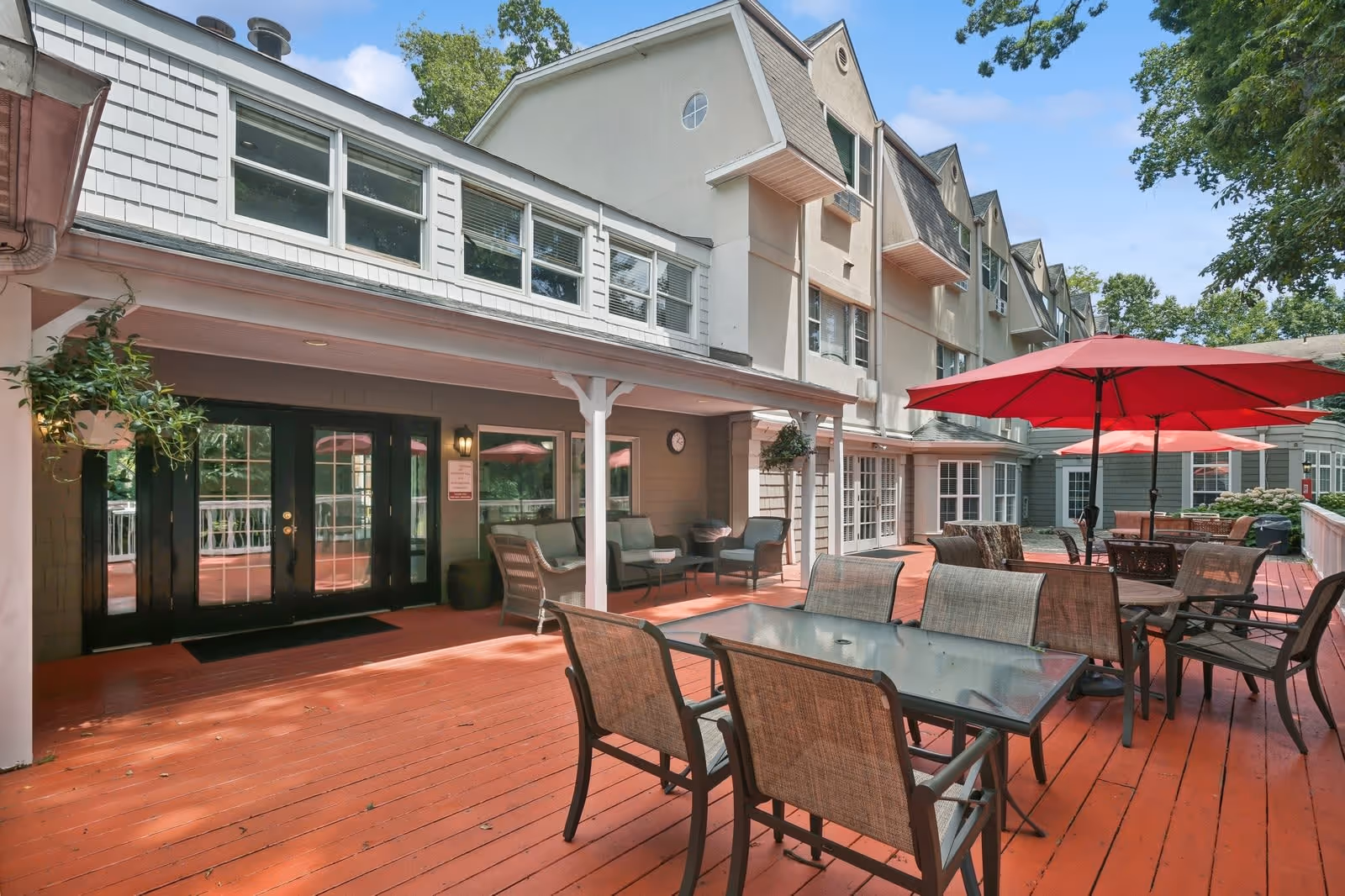 Outdoor patio area at The Terrace at Chestnut Hill featuring multiple tables with chairs and large red umbrellas, adjacent to a multi-story building with large windows and doors leading inside. The patio has a wooden deck floor and some greenery is visible around the area.
