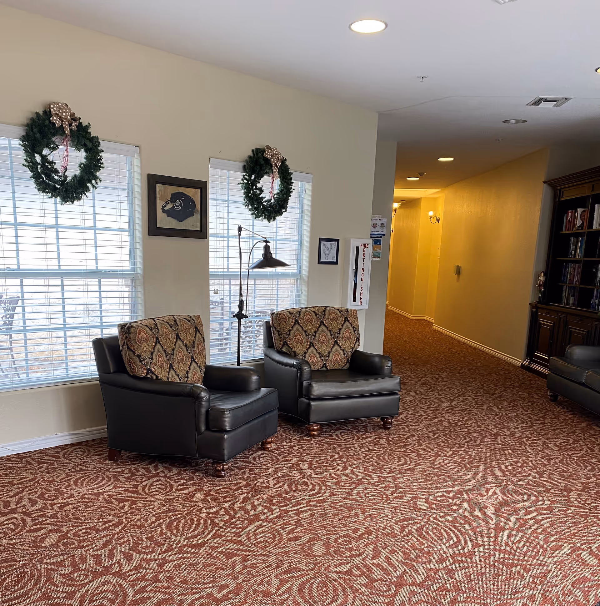 A cozy interior seating area with two leather armchairs featuring patterned cushions, positioned in front of two windows decorated with holiday wreaths. A floor lamp stands between the chairs, and a framed picture hangs on the wall. The carpet has a red and beige floral pattern, and a hallway extends into the background with warm lighting. A wooden bookshelf is partially visible on the right side.