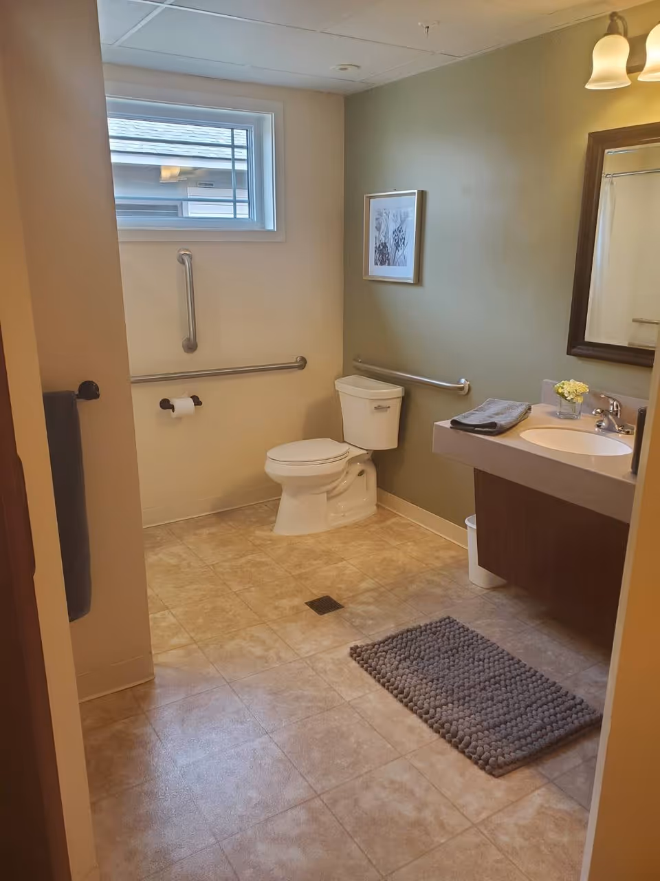 A clean, accessible bathroom with beige tiled floor, a white toilet with grab bars on the walls, a sink with a countertop and mirror above it, a small window, a gray bath mat on the floor, and a framed picture on the wall.