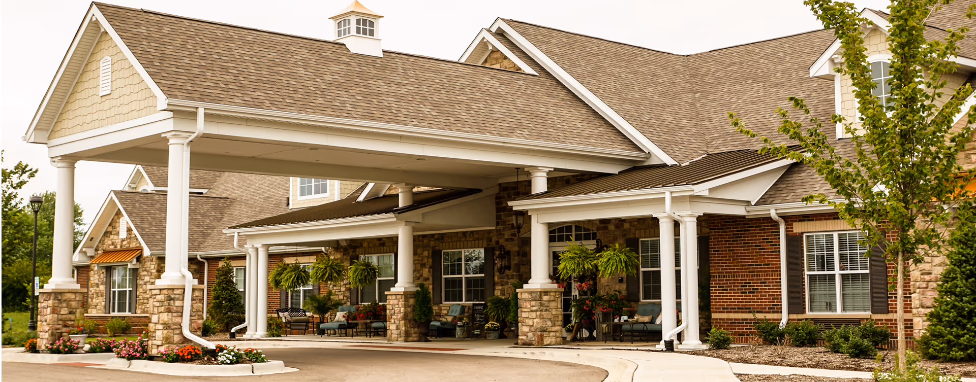 Front exterior view of a senior living facility with a covered entrance supported by white columns. The building features a combination of brick and stone walls, multiple windows with shutters, hanging plants, and outdoor seating areas with chairs and small tables. There are landscaped flower beds and a tree near the entrance.