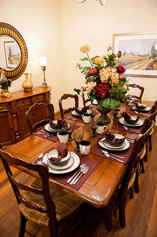 A wooden dining table set for six with matching chairs, each place setting includes a plate, bowl, cup, and silverware on a burgundy placemat. A large floral centerpiece with red, pink, and yellow flowers is in the middle of the table. In the background, there is a wooden sideboard with decorative items and a round mirror above it, as well as a framed landscape painting on the wall.