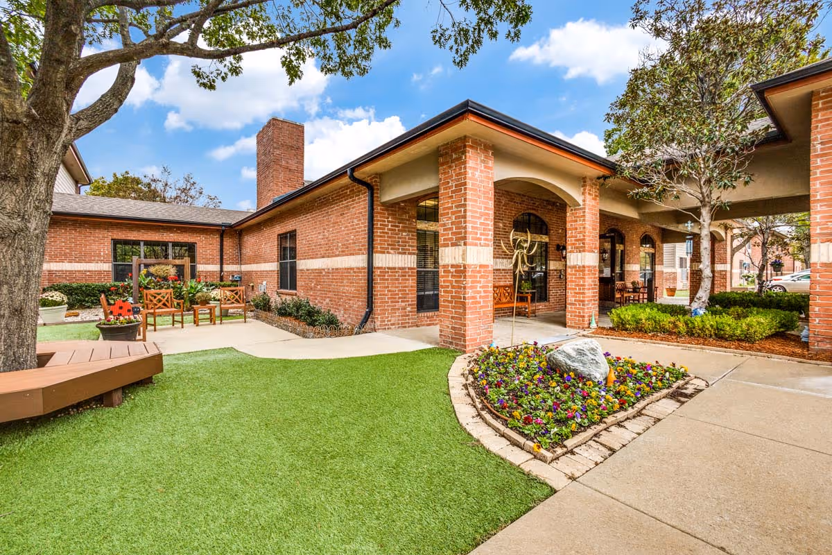 Outdoor courtyard area of a senior living facility with brick buildings, a flower bed with colorful flowers and a large rock, green artificial turf, a tree with a circular wooden bench around its base, and wooden chairs arranged near the building under a covered walkway.