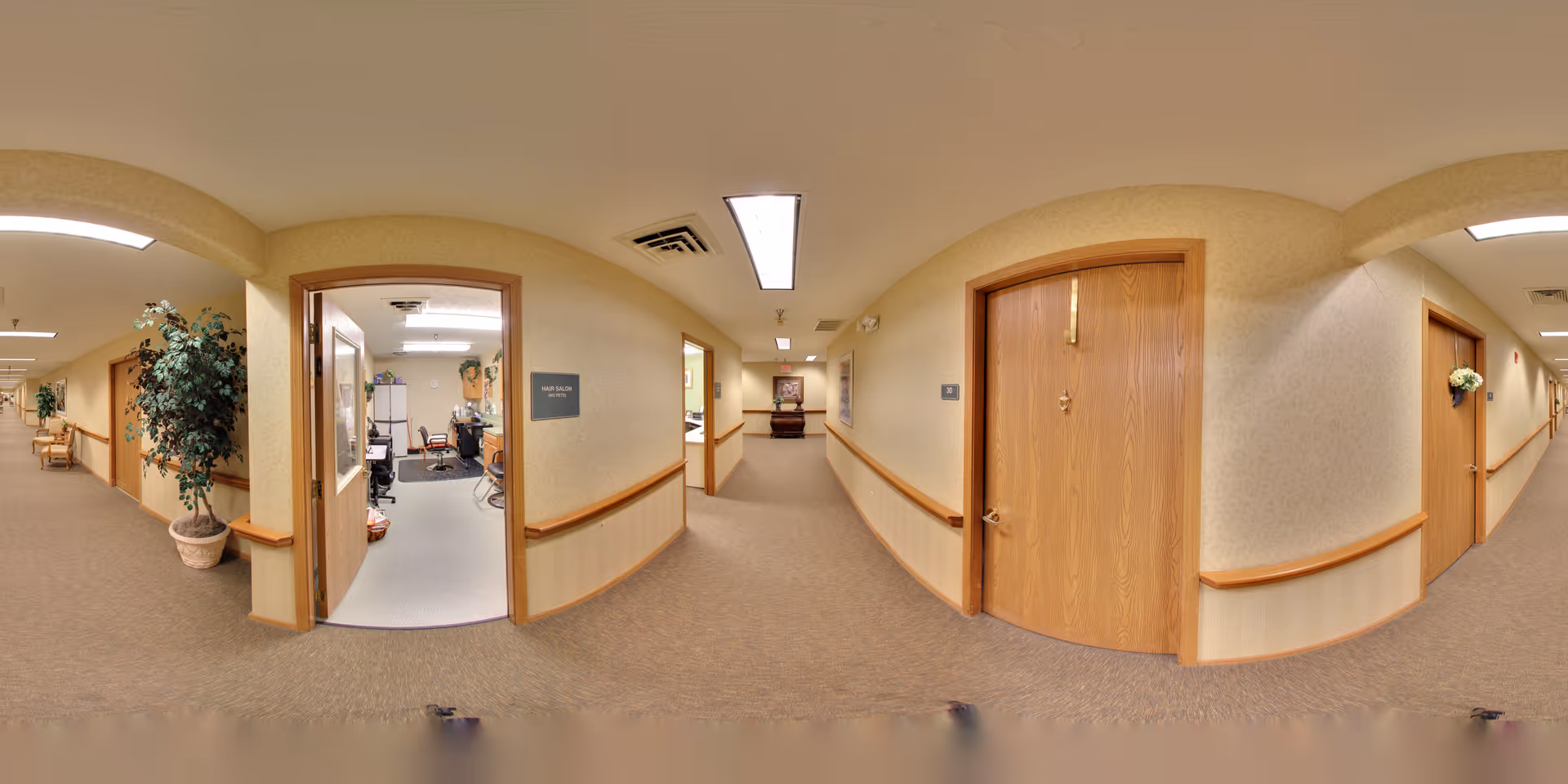 A wide-angle view of a hallway in a senior living facility with beige walls and carpeted floors. Several wooden doors line the hallway, one of which is open revealing an office with chairs, a desk, and office equipment. There are handrails along the walls and a potted plant near the open door. The ceiling has fluorescent lighting panels.