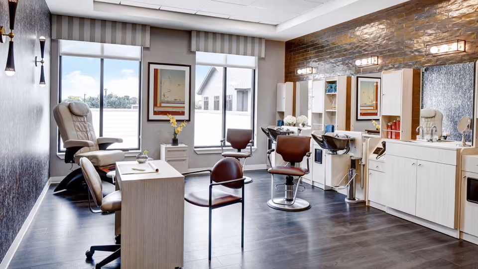 A bright and modern salon area with large windows, featuring salon chairs, hair washing stations, a manicure table, and a comfortable reclining chair. The room has wood flooring, white cabinetry, and decorative lighting fixtures on the walls.