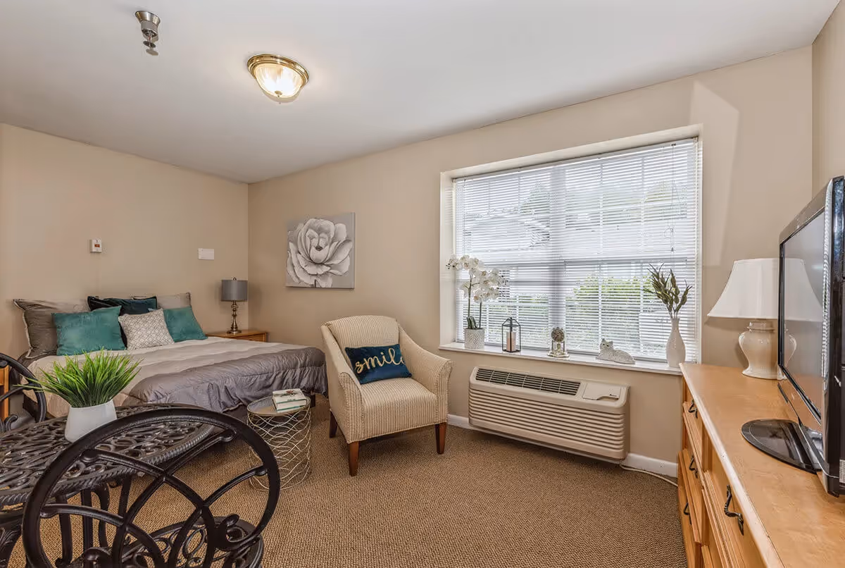 Furnished studio bedroom with a bed, upholstered chair, window with blinds, TV on a dresser, and a small table.