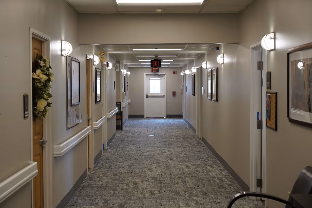A well-lit hallway in a senior living facility with multiple doors on both sides, wall-mounted lights, framed pictures, and a carpeted floor leading to an exit door at the end.