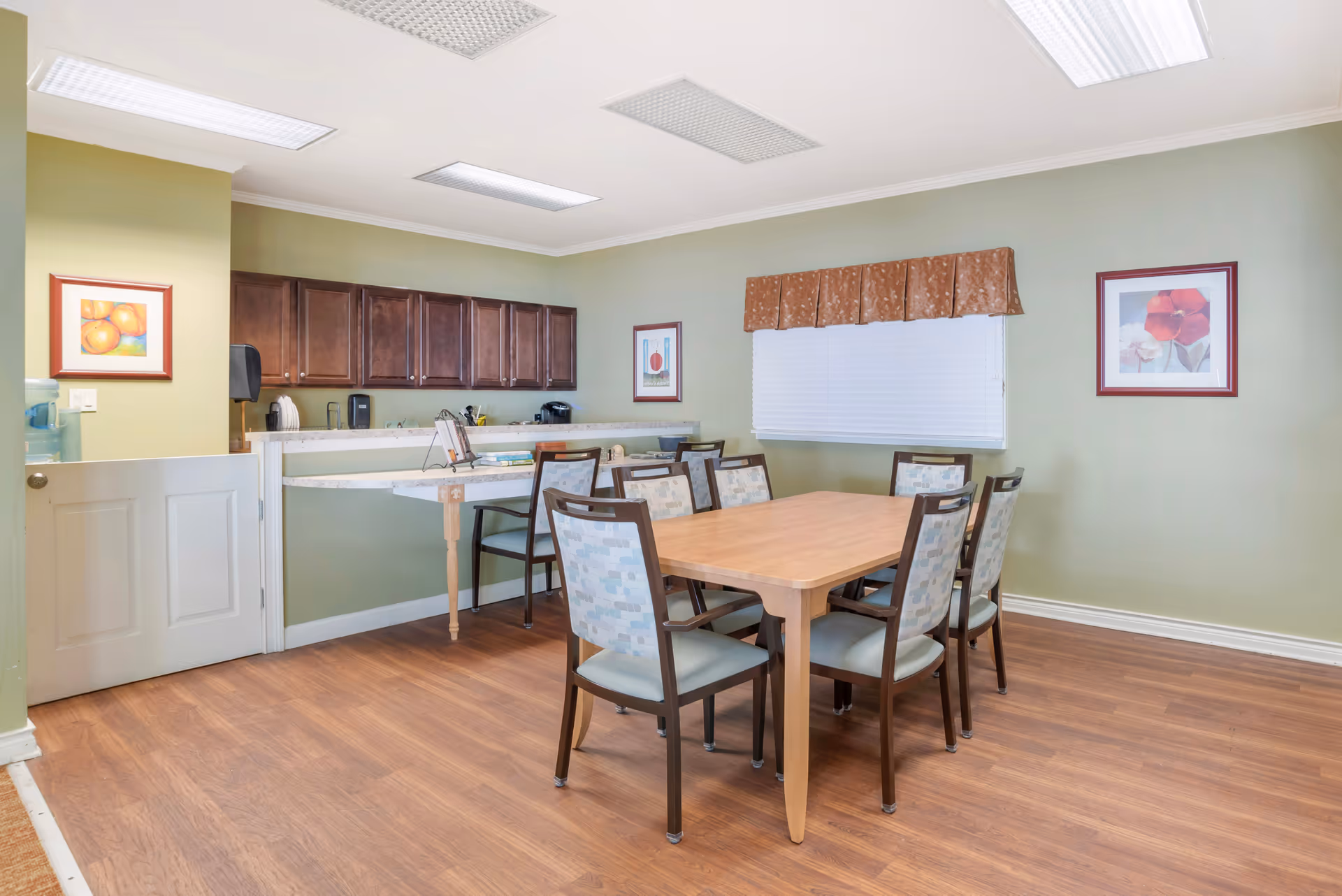 A dining area with a wooden table and six chairs with patterned upholstery. The room has light green walls, wood flooring, and a window with closed white blinds and a brown valance. There are three framed pictures on the walls and a counter with dark wooden cabinets and various kitchen items in the background.