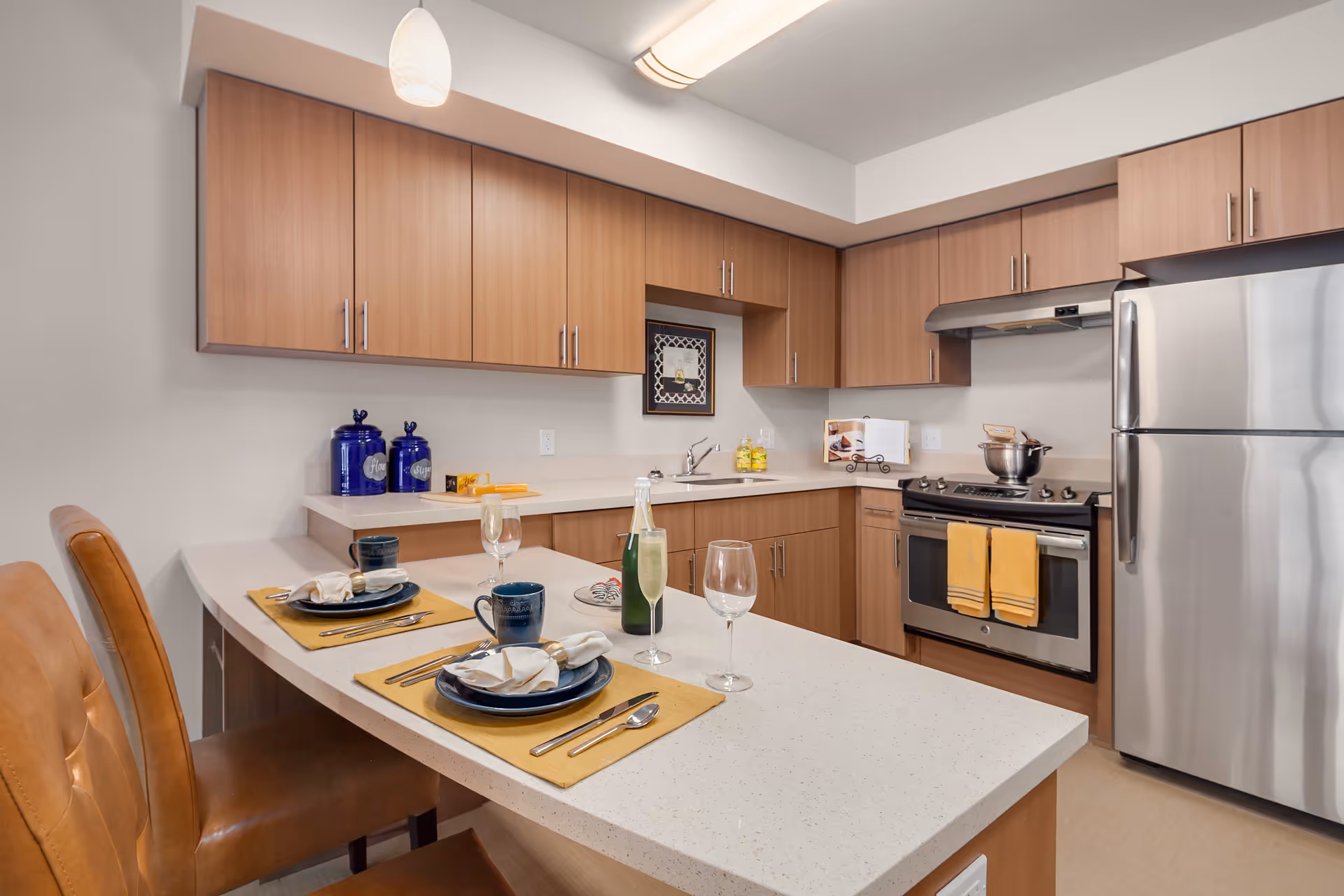 Modern kitchen with wooden cabinets, stainless steel refrigerator and stove, a white countertop island with two place settings including plates, napkins, utensils, mugs, and wine glasses. Two brown leather chairs are positioned at the island.