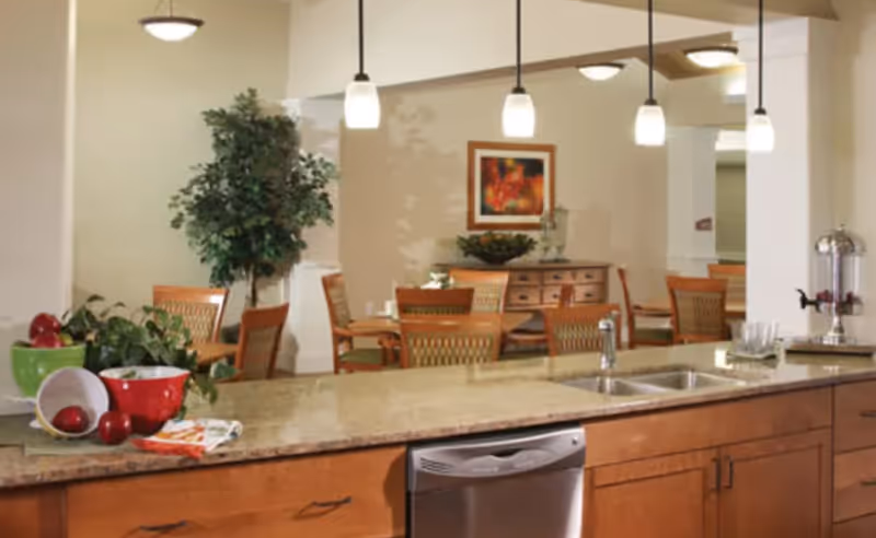 Interior view of a dining area in a senior living facility with wooden chairs and tables, a sideboard with decorative items, and a kitchen counter with a sink, dishwasher, and bowls of fruit. Pendant lights hang above the counter, and there is a large potted plant near the dining tables.