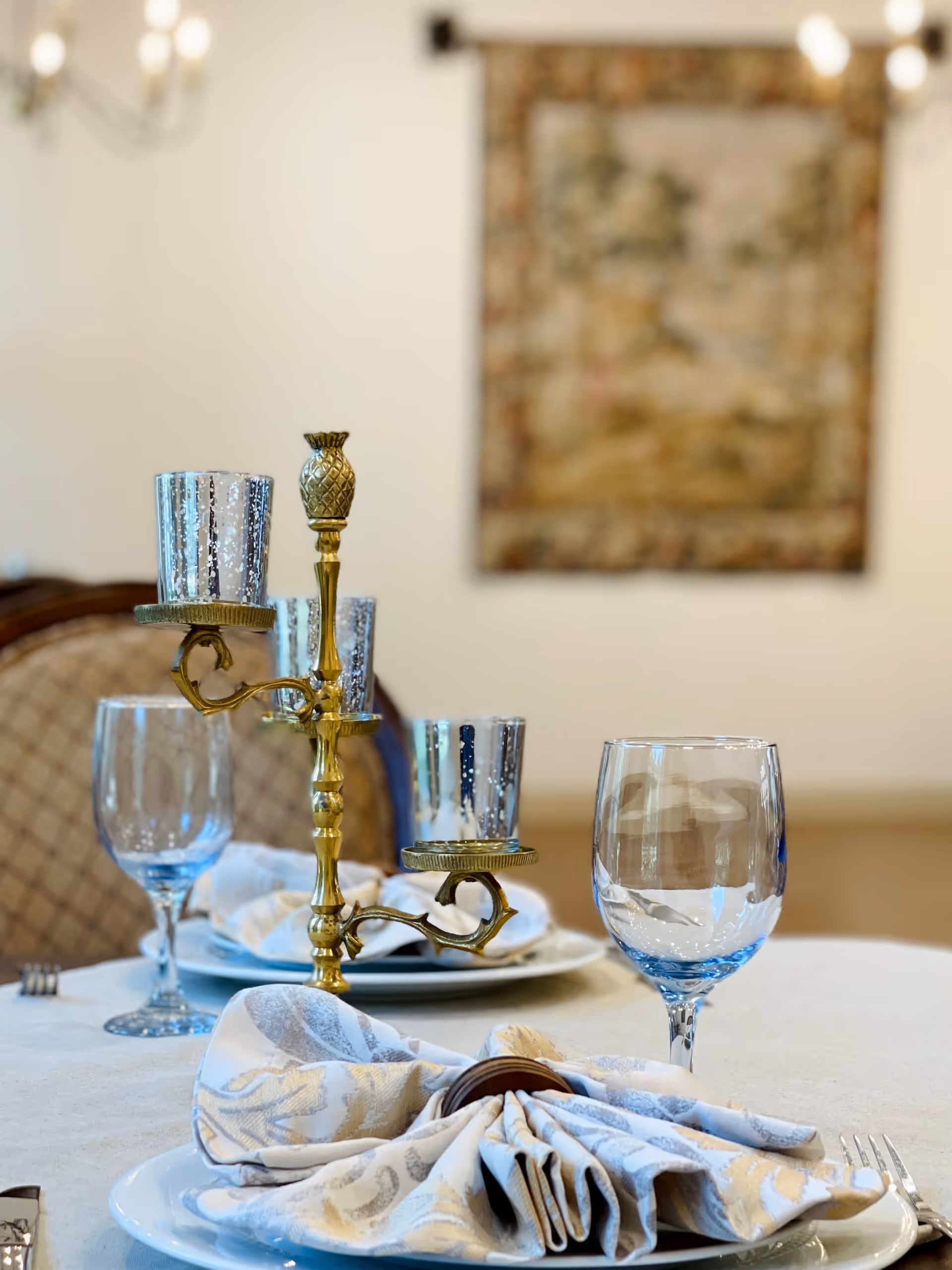 Close-up of a dining table set with a decorative brass candle holder, two empty wine glasses, and a folded patterned cloth napkin on a white plate. In the background, there is a blurred upholstered chair and a framed artwork on the wall.
