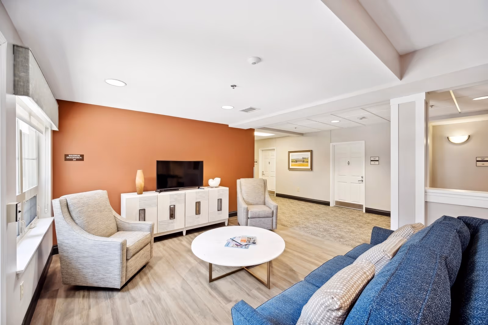 A bright and modern living room area with a blue sofa, two gray armchairs, a round white coffee table with magazines, a white TV console with a flat-screen TV, and a burnt orange accent wall. The room has wood flooring and a window with a gray valance letting in natural light.