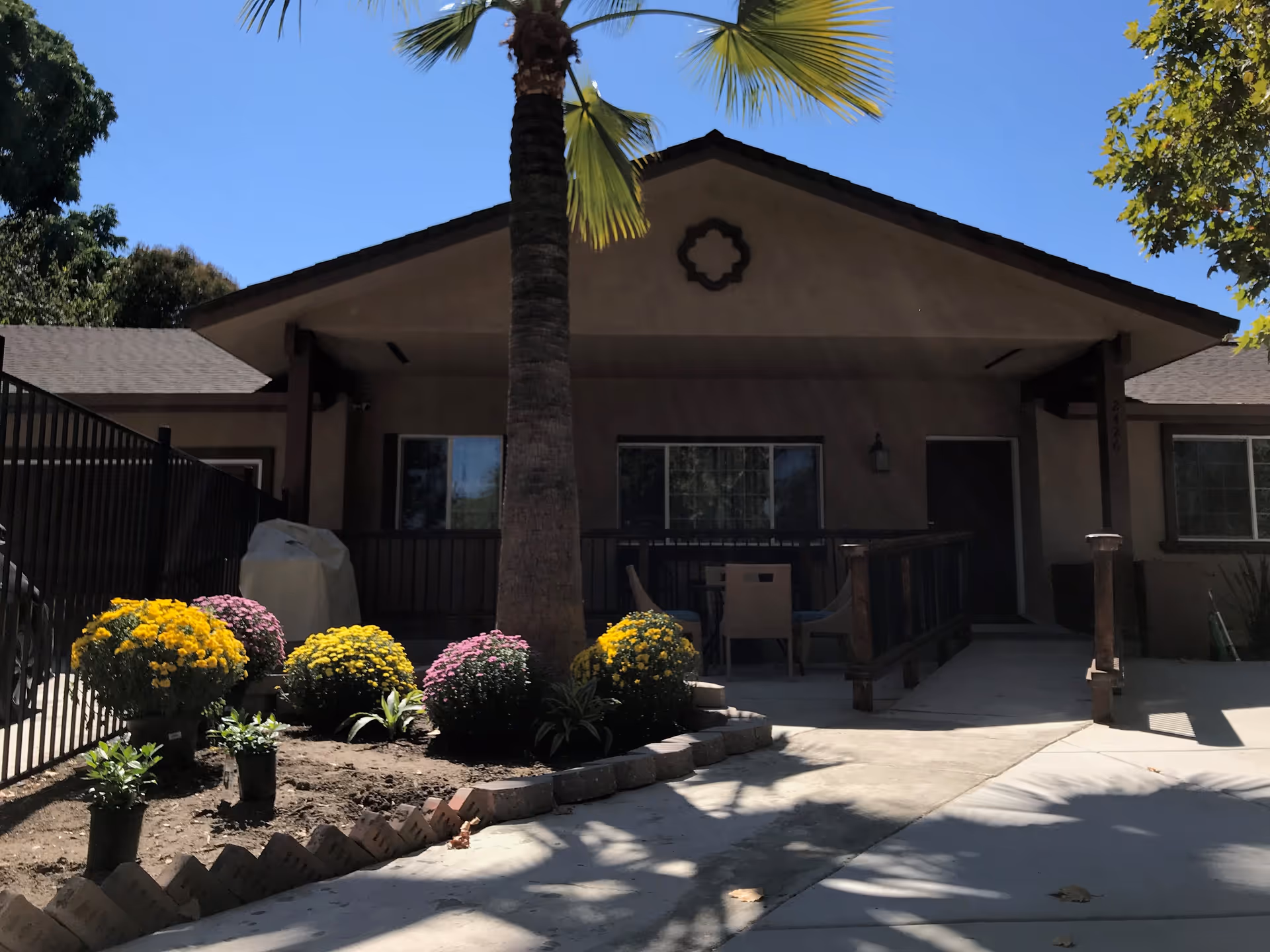 Front exterior view of a single-story building with a covered porch, a palm tree in the foreground, and colorful potted flowers along the walkway under a clear blue sky.