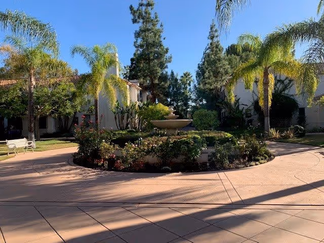 A sunny outdoor courtyard with a circular paved area surrounding a central fountain. The courtyard is landscaped with palm trees, bushes, and flowering plants. Two buildings with light-colored walls and red-tiled roofs are visible in the background, along with a white bench on the left side.