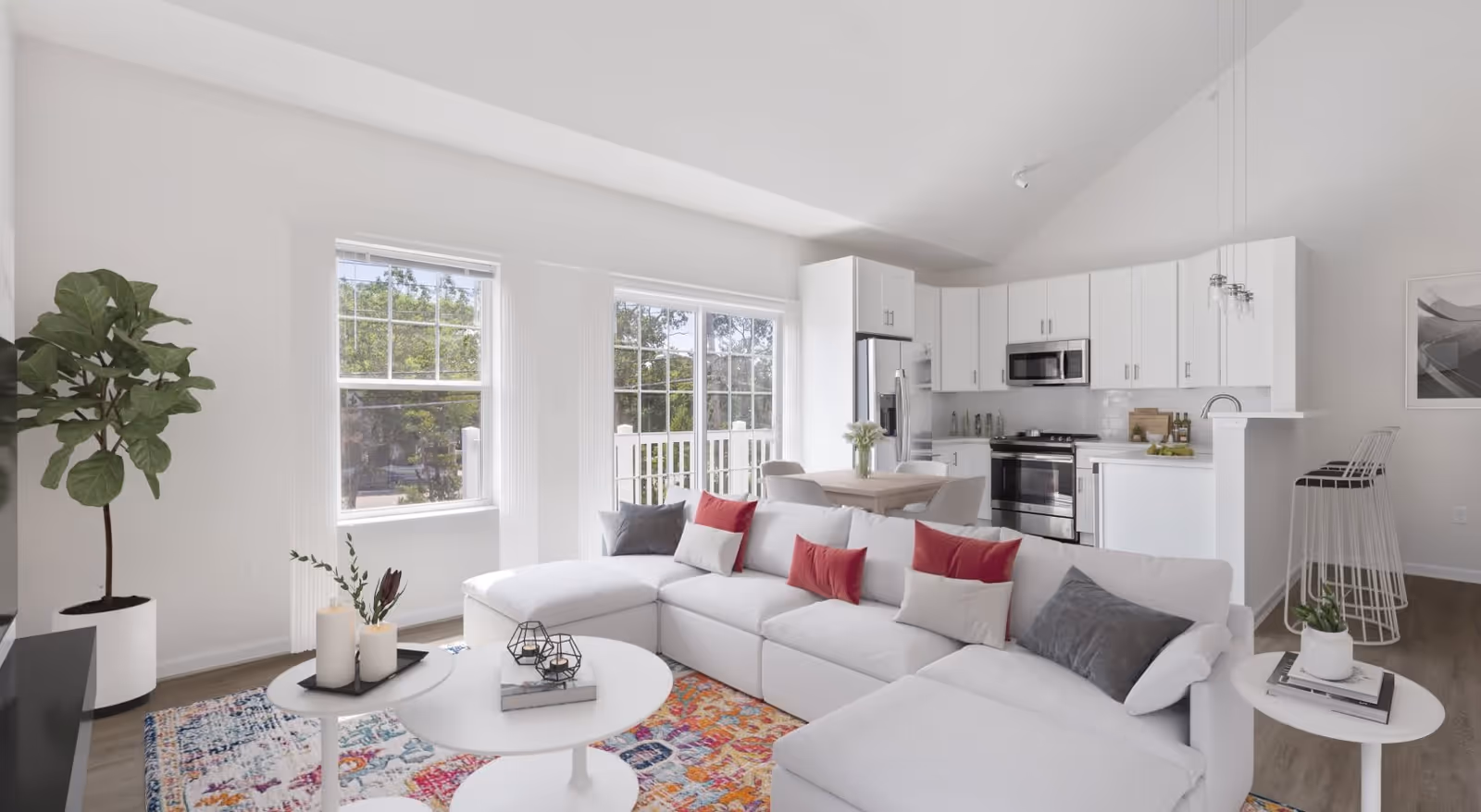 Bright and modern open-concept living room and kitchen area with white walls and ceiling, large windows letting in natural light, a white sectional sofa with red and gray pillows, colorful area rug, white round coffee tables with decorative items, a tall green potted plant, a dining table with chairs, and a kitchen with white cabinets, stainless steel appliances, and a breakfast bar with stools.