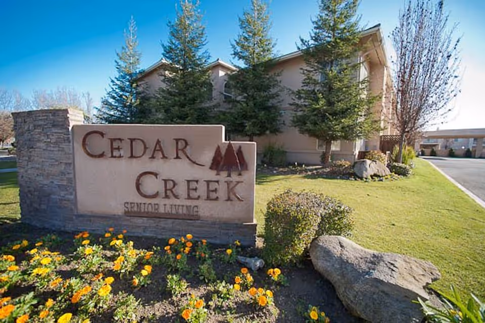 Entrance stone sign reading 'Cedar Creek Senior Living' with landscaped flowers, trees, and the facility building in the background.
