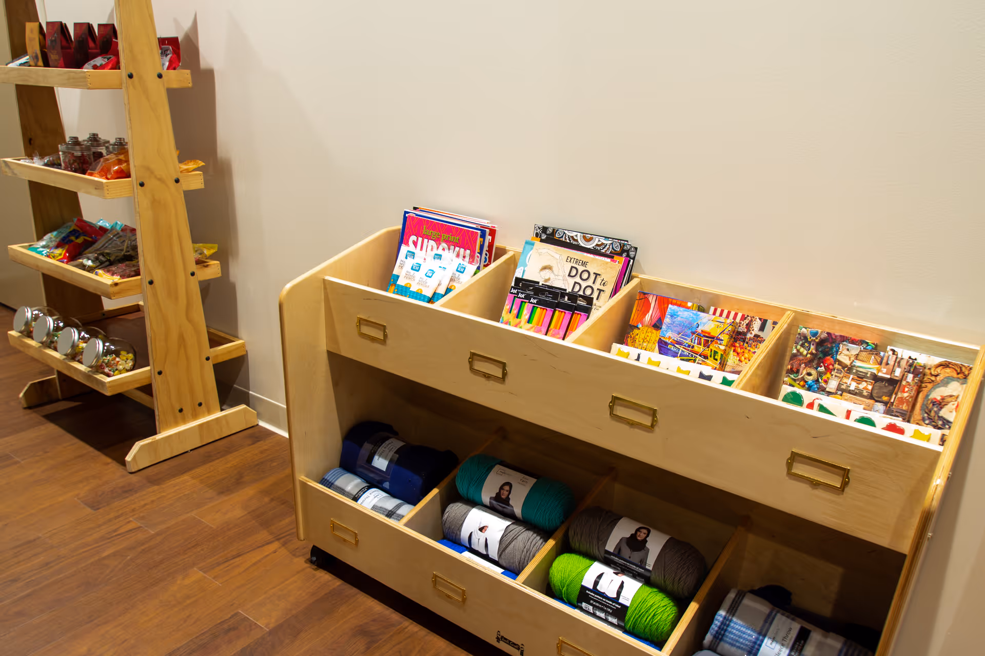 A wooden storage unit with compartments holding yarn, magazines, coloring books, and art supplies, next to a wooden shelf stocked with various snacks and jars, all set against a beige wall and wooden floor.