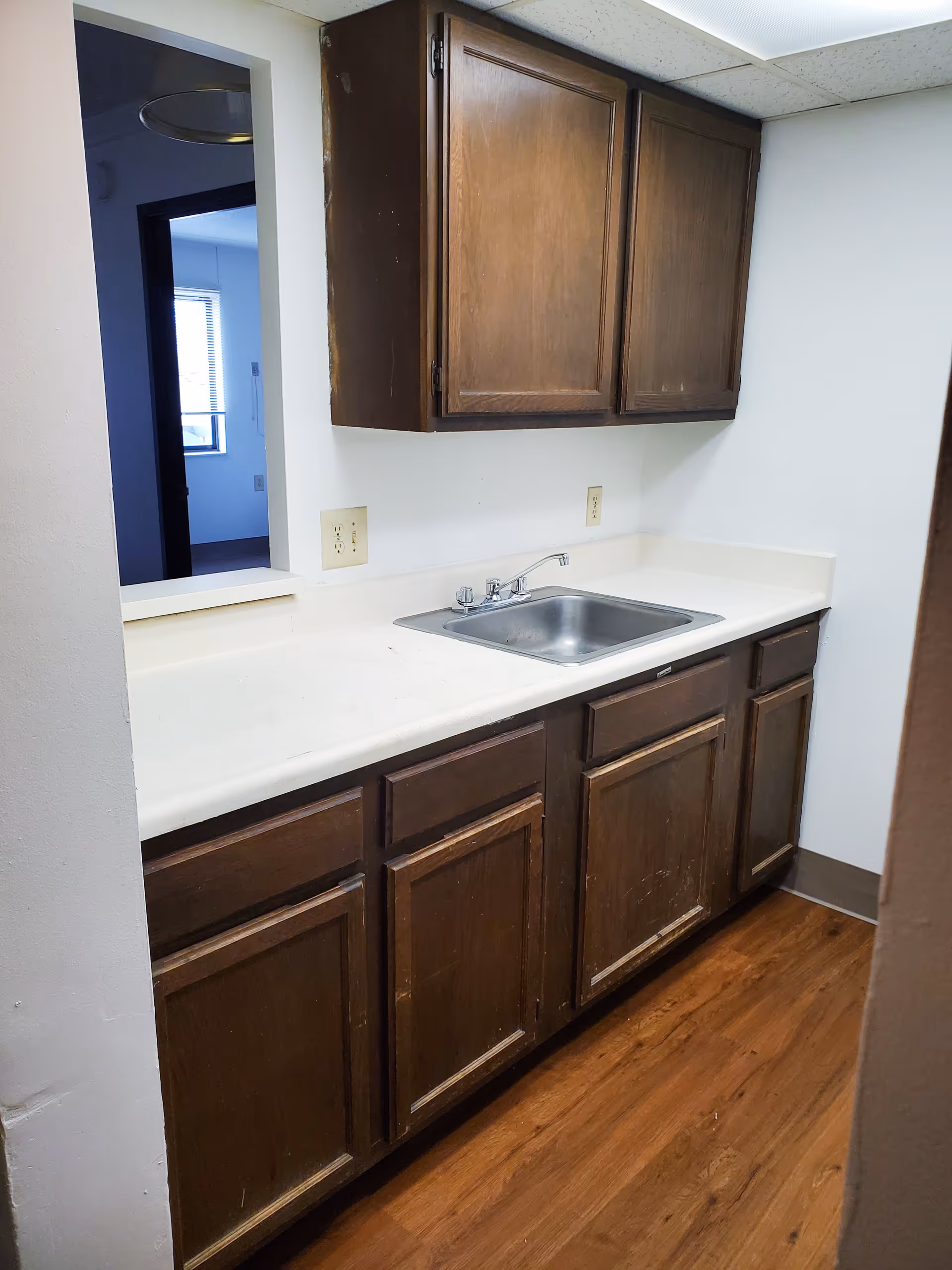 Small kitchen area with wooden cabinets above and below a white countertop. A stainless steel sink with a faucet is installed in the countertop. The floor is wood-patterned, and there is a pass-through window opening to another room with a window visible in the background.