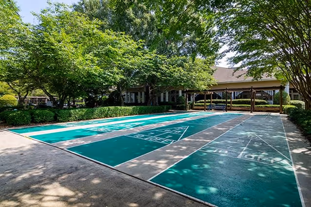 Outdoor shuffleboard courts surrounded by trees and bushes with a building in the background under a clear sky.