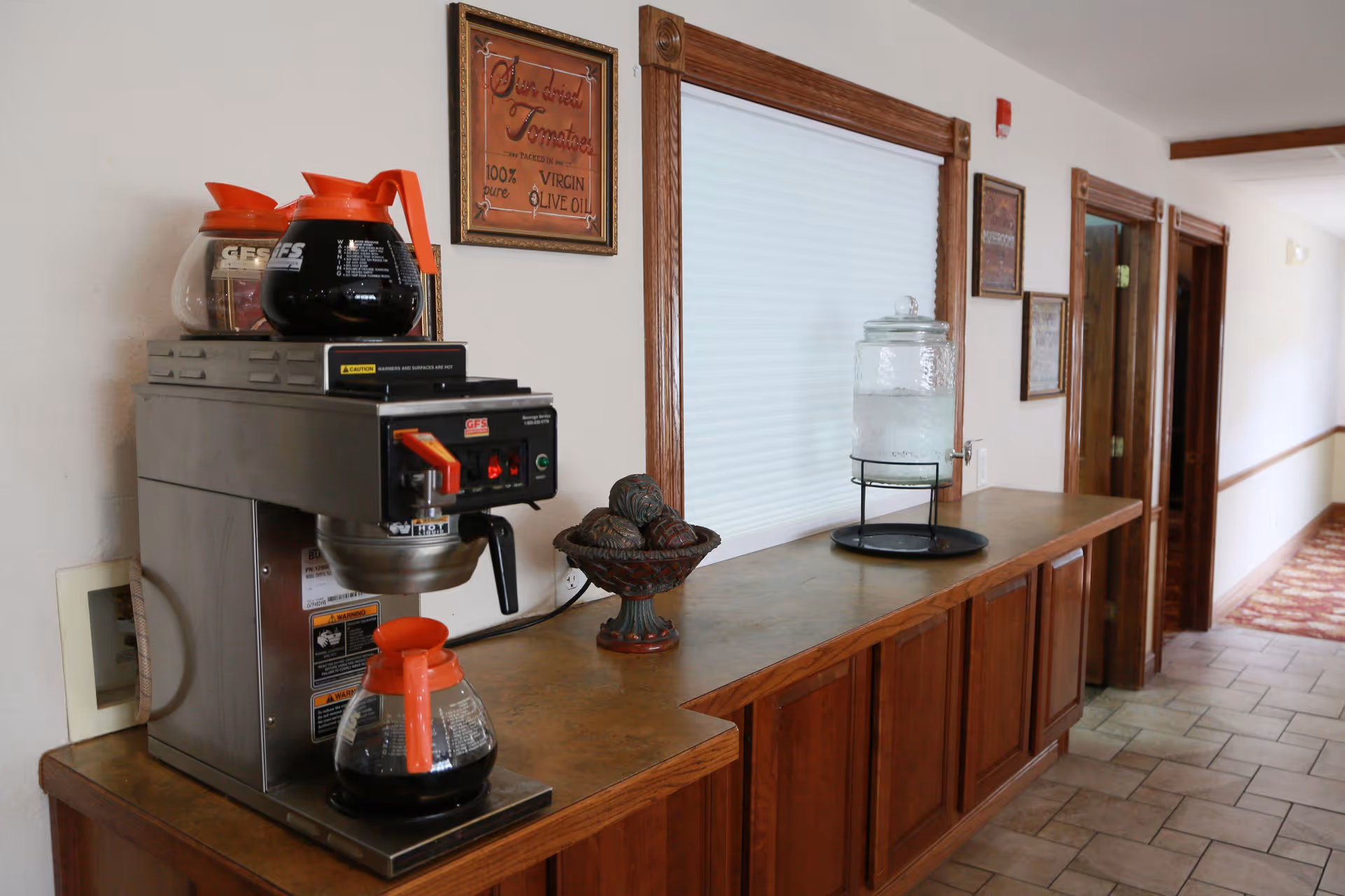 Coffee station with coffee maker, coffee carafes, and a glass water dispenser on a wooden counter in a hallway.