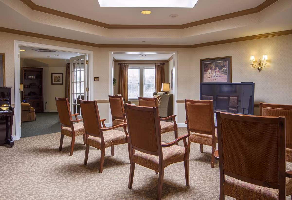 A well-lit common area with several wooden chairs arranged in rows facing a large flat-screen TV mounted on the wall. The room has beige walls with a framed painting and wall sconces providing warm lighting. There is a window with curtains in the background and an adjacent room visible through an open doorway.