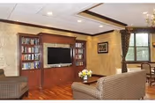 Communal living room with a built-in entertainment center and TV, upholstered chairs, a coffee table with flowers, and a dining area by a window.