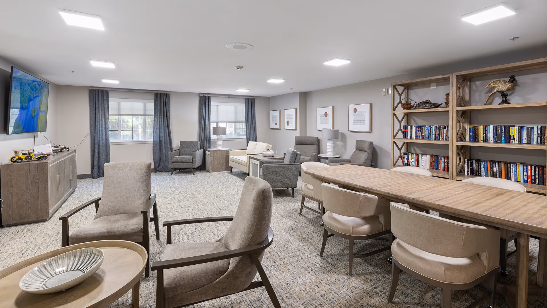 A spacious and well-lit common area with multiple seating options including armchairs and a long wooden table with chairs. There are two windows with dark curtains, a large bookshelf filled with books and decorative items, a wall-mounted TV, and framed artwork on the walls. The room has a neutral color palette with beige and gray tones.