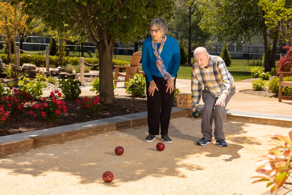 An elderly man and woman playing bocce ball outdoors on a sandy court surrounded by greenery and flowers. The man is in the process of throwing a bocce ball while the woman watches attentively. There are trees, bushes, and benches in the background.