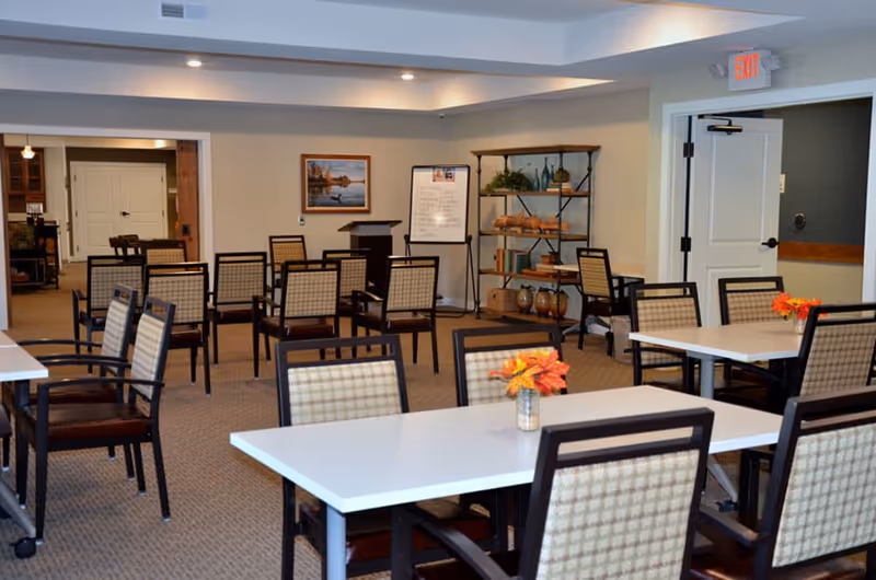 A well-lit dining room in a senior living facility with multiple tables and chairs arranged neatly. Each table has a small vase with orange and yellow flowers. In the background, there is a bookshelf with decorative items, a framed picture on the wall, and a whiteboard on a stand. The room has beige walls and carpeted floors, with an exit door visible on the right side.