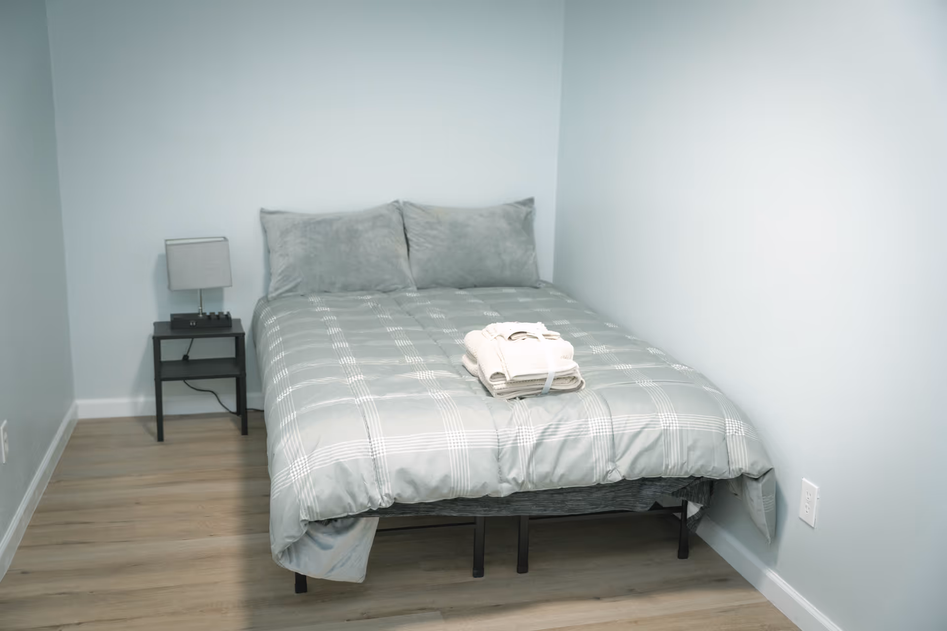 A small bedroom with light blue walls and wooden flooring featuring a bed with a gray plaid comforter and two matching pillows. A neatly folded set of towels is placed on the bed. Next to the bed is a black nightstand with a small gray lamp and an electronic device on it.