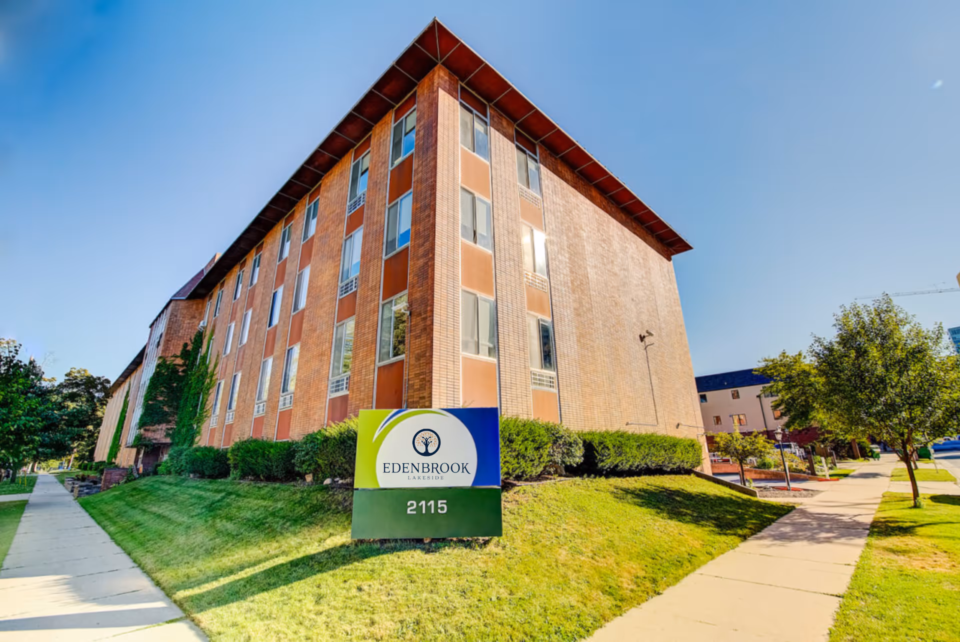 Brick multi-story senior living building with a lawn and an 'Edenbrook Lakeside' sign reading 2115 by the sidewalk.