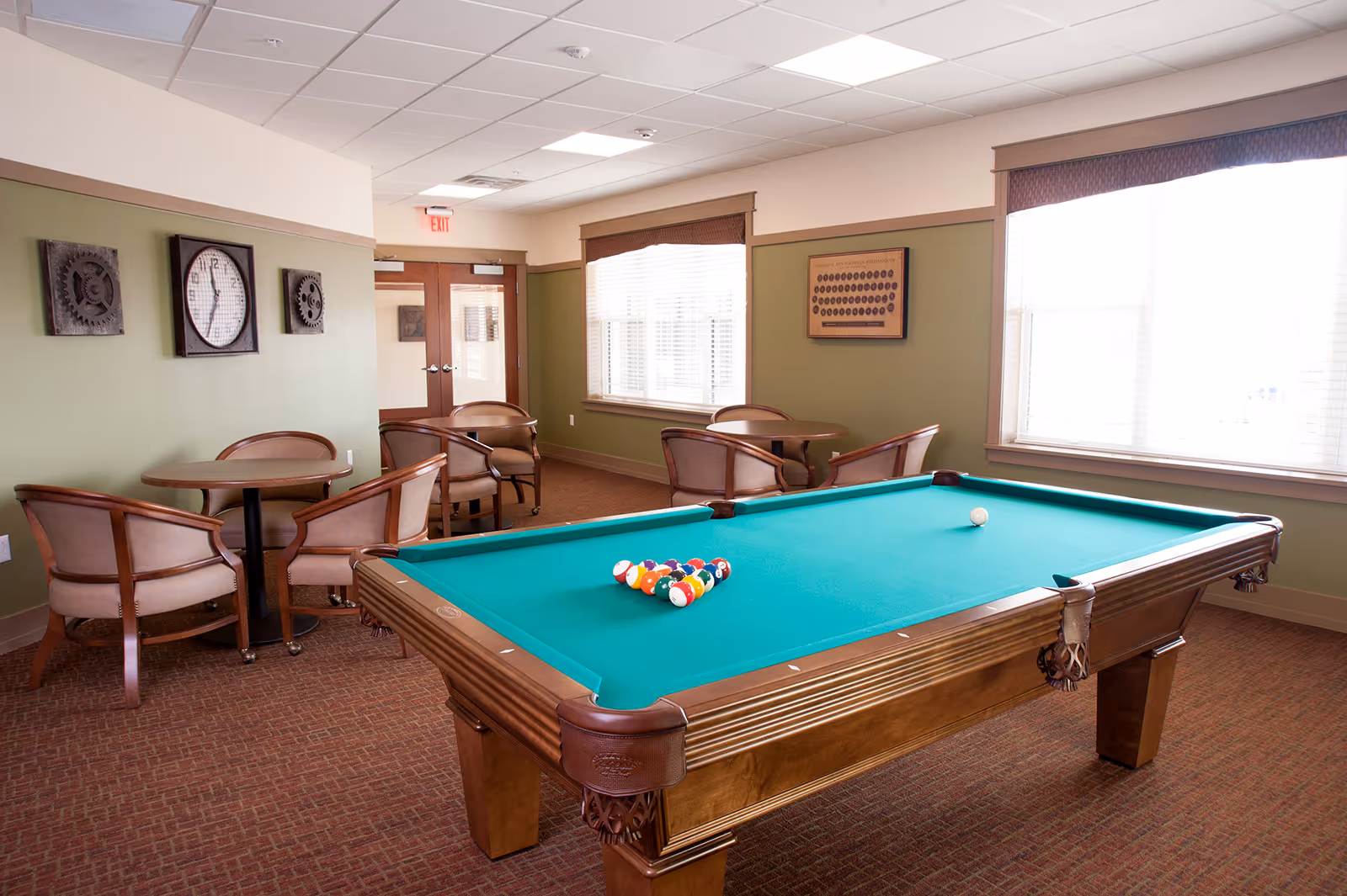 A well-lit recreational room with a pool table in the center, set up for a game with billiard balls arranged in a triangle and a cue ball nearby. The room has green walls with wooden trim, two large windows with blinds, and several round tables with cushioned chairs arranged around the room. Decorative wall art and a clock are mounted on the walls.