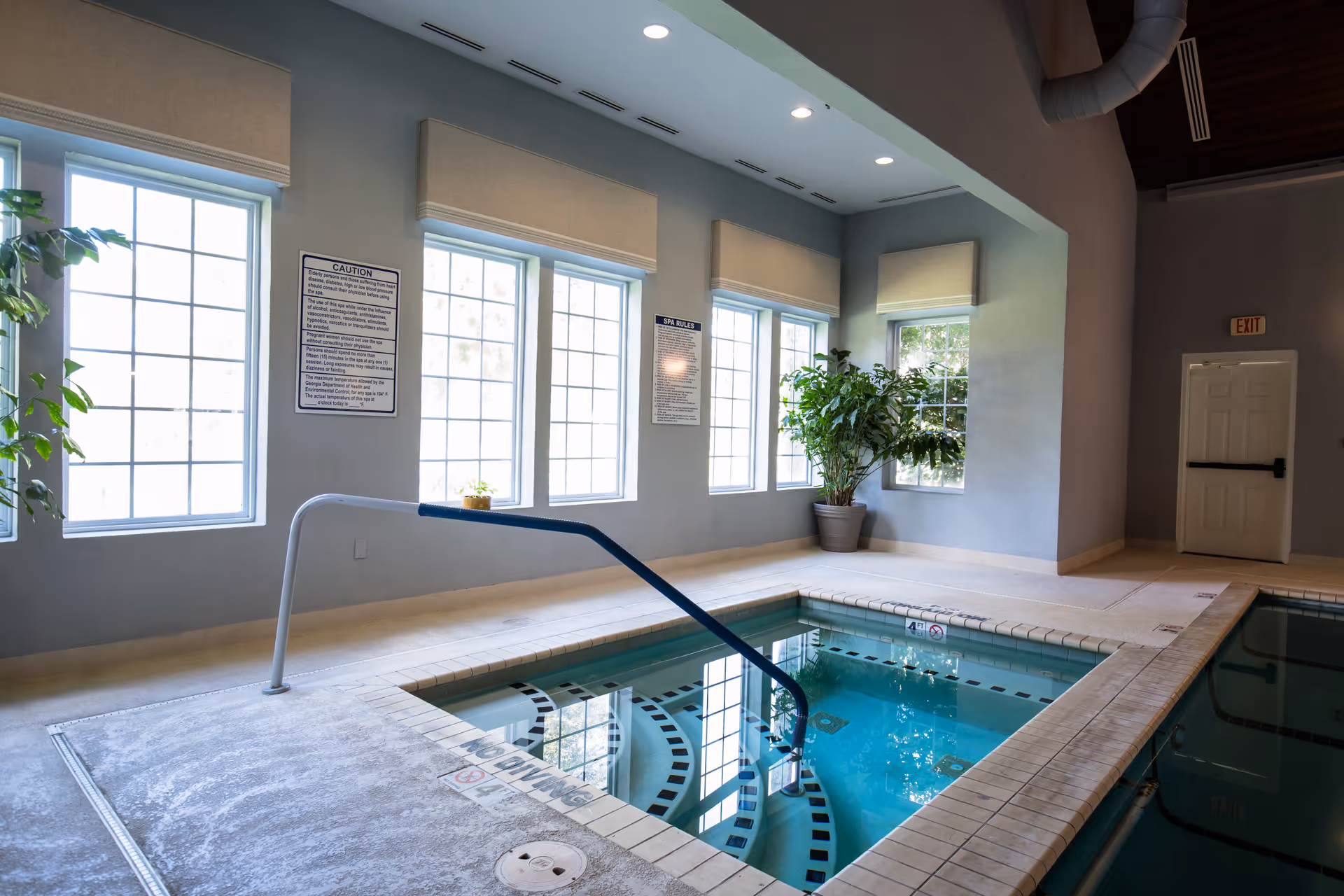 Indoor pool area with a small square hot tub in the foreground and a larger pool partially visible to the right. The room has large windows with beige shades, potted plants, and safety signs on the wall. There is a metal handrail leading into the hot tub and an exit door in the background.