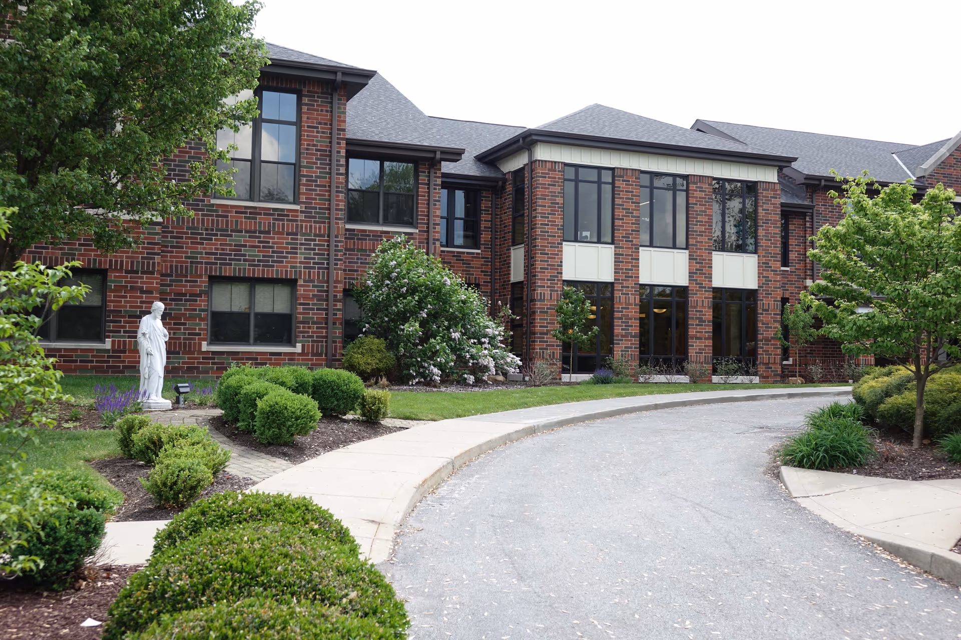 Curved driveway leading to the entrance of a two-story brick building with large windows, surrounded by well-maintained landscaping including bushes, trees, and a white statue near the left side.