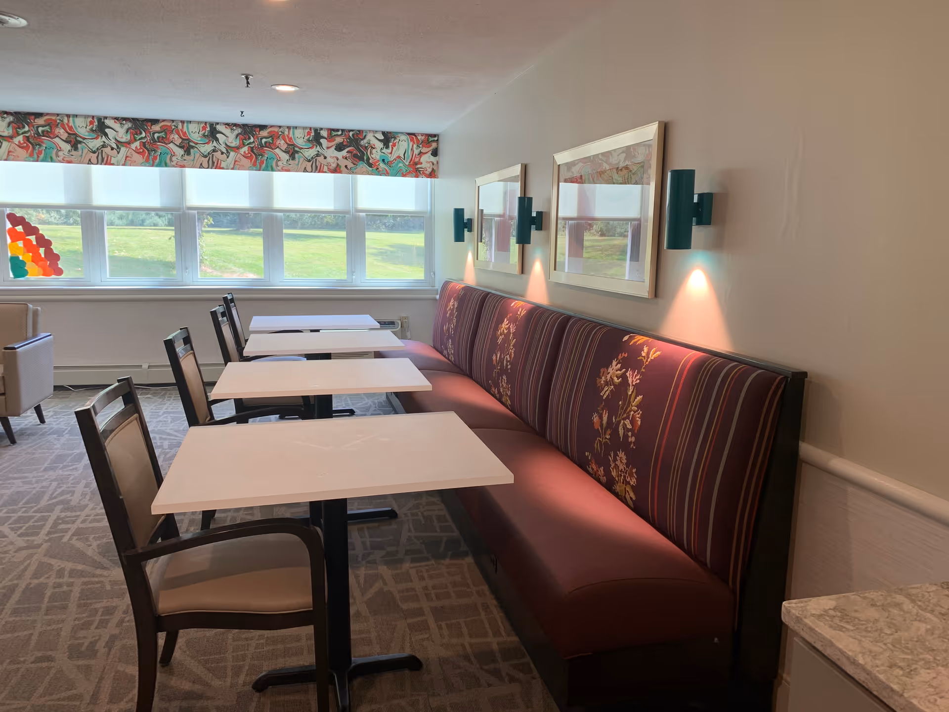 A dining area with three white tables aligned in a row, each paired with a wooden chair on one side and a long upholstered bench with floral and striped patterns on the other side. The room has a large window with a colorful valance and a view of green grass outside. Two framed pictures and wall-mounted lights are above the bench seating.