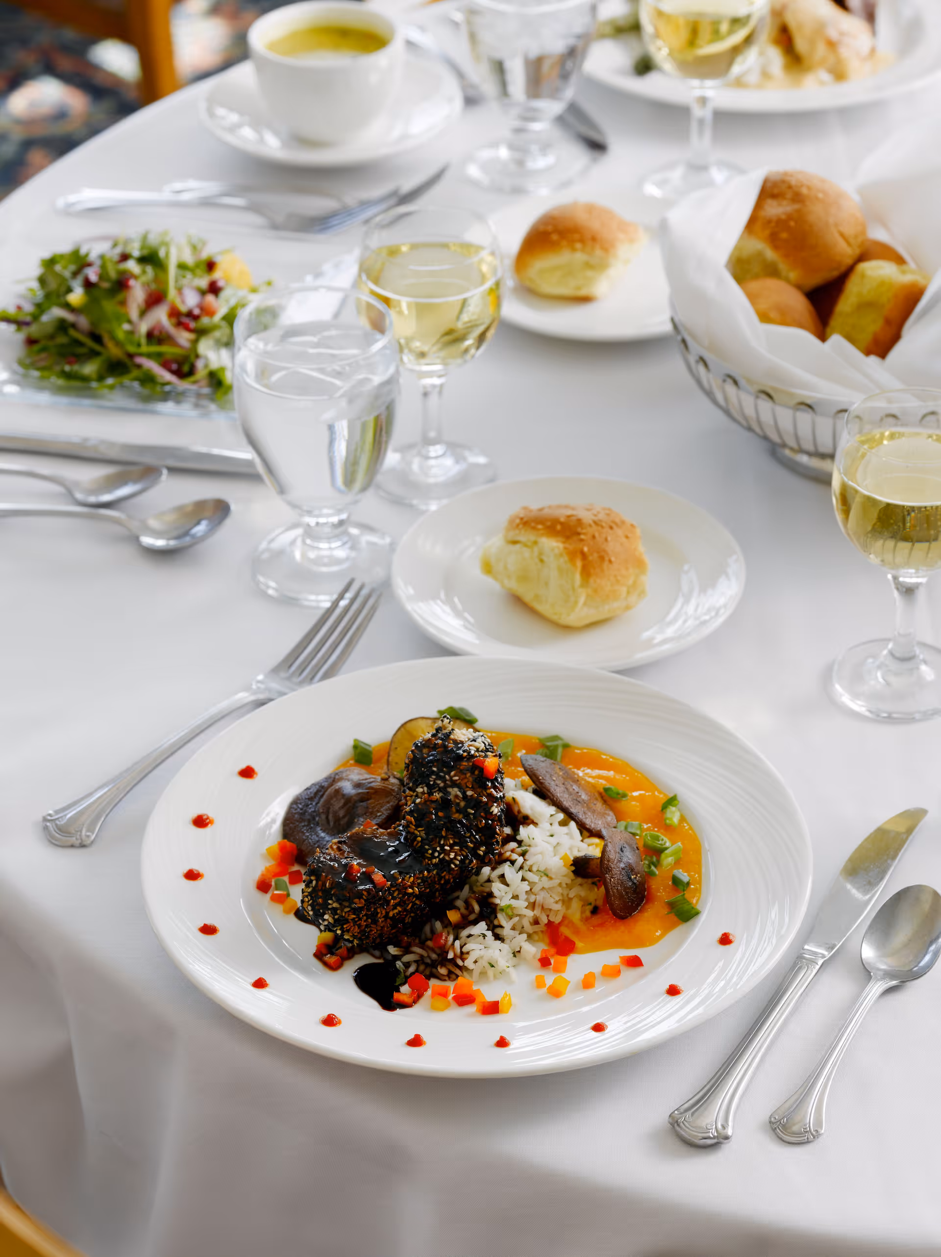 A white tablecloth-covered dining table set with a gourmet meal including a plate of rice, vegetables, and a dark sauce-covered main dish, a small bread roll on a side plate, a basket of bread rolls, a glass of white wine, a glass of water, and a bowl of green soup. Silverware is neatly arranged around the plates.