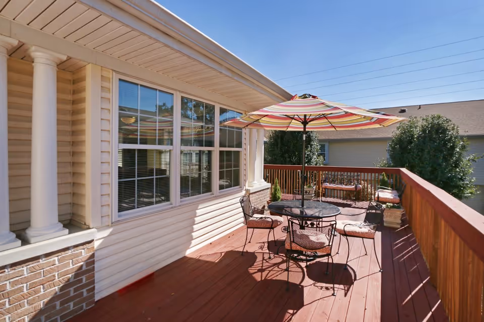 Outdoor wooden deck attached to a building with beige siding and white columns. The deck has a round metal table with four cushioned chairs and a large striped umbrella providing shade. There are potted plants and a bench with cushions along the wooden railing. The sky is clear and blue.