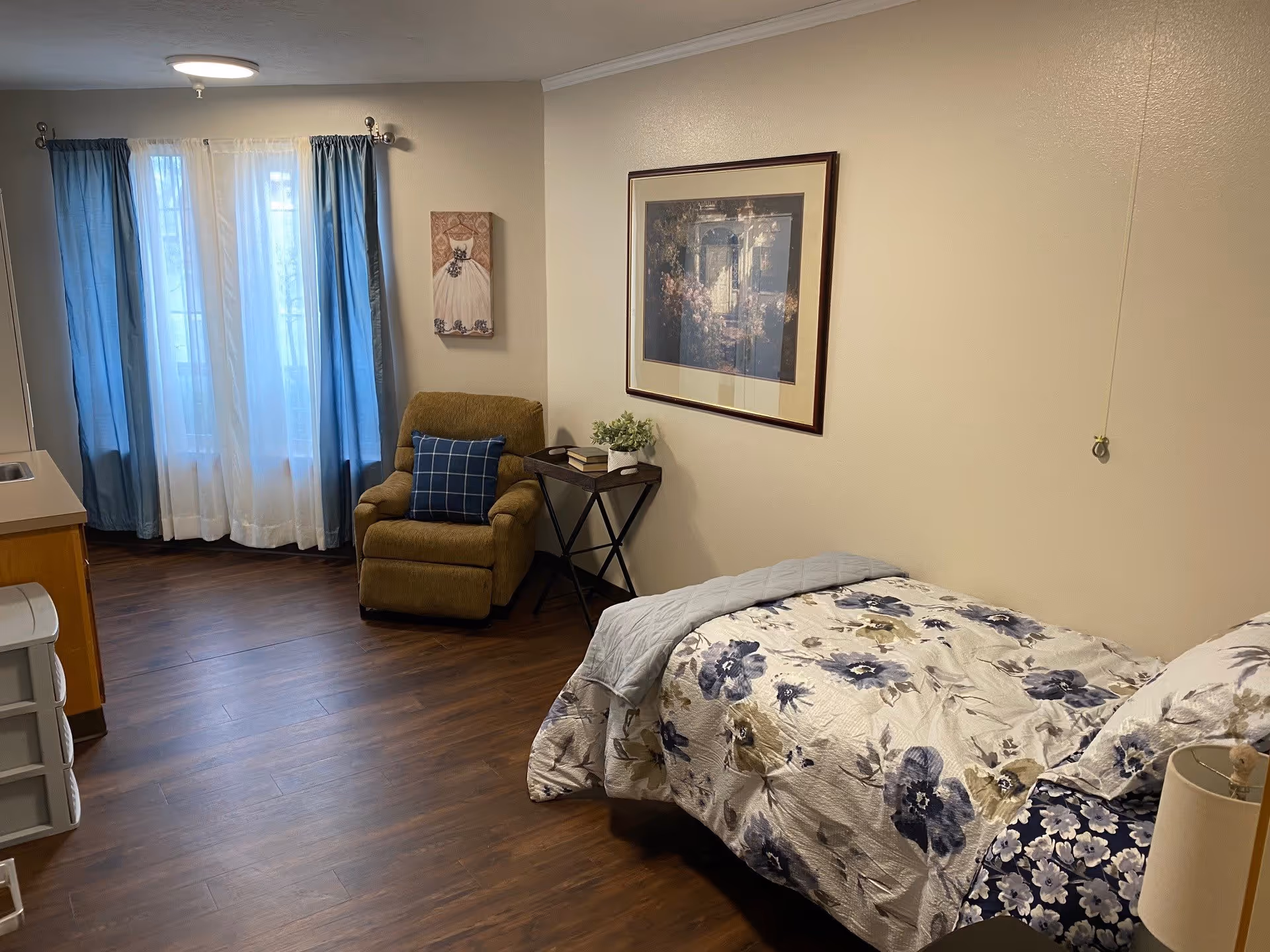 A cozy bedroom in a senior living facility featuring a single bed with floral bedding, a beige recliner chair with a blue checkered pillow, a small side table with books and a plant, wooden flooring, and a window with blue and white curtains. The walls are decorated with framed artwork.