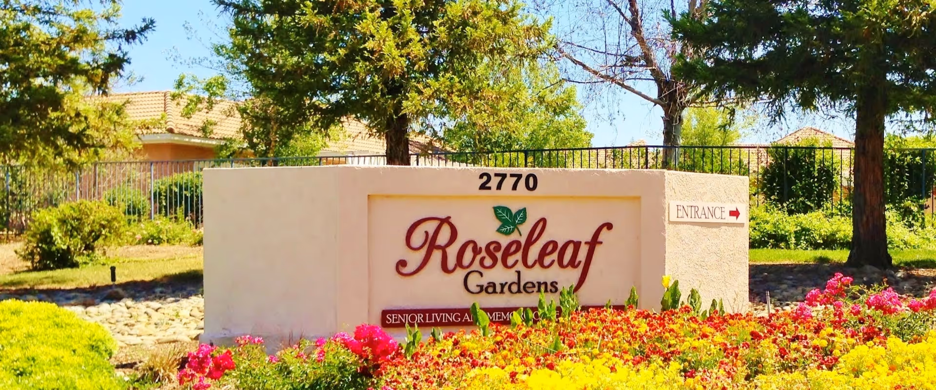 Outdoor view of a sign for Roseleaf Gardens senior living and memory care facility, surrounded by colorful flowers and greenery with trees and houses in the background.