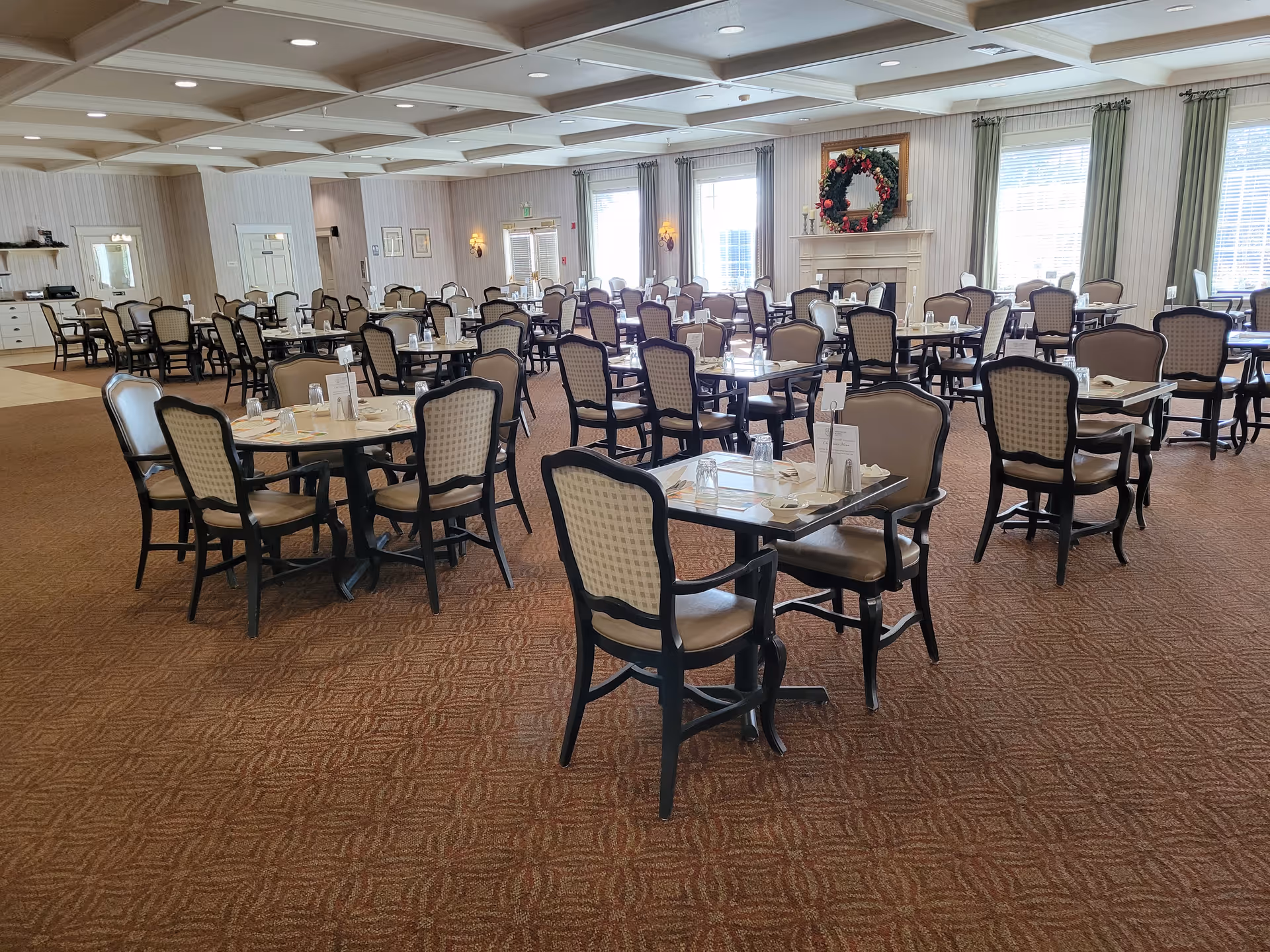 Large dining room with multiple set tables and upholstered chairs in a senior living facility.