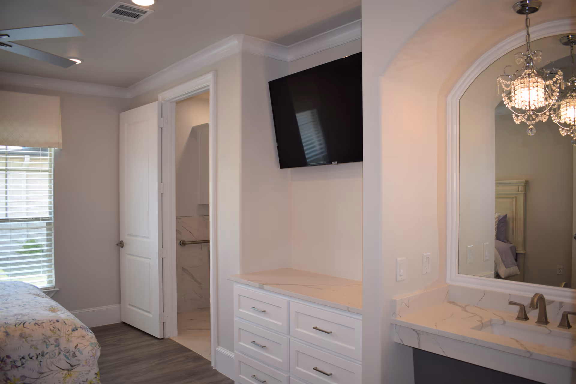 A bedroom with a floral bedspread partially visible on the left, a window with blinds, a white door leading to a bathroom with marble walls and a grab bar, a wall-mounted flat screen TV above a white dresser with six drawers, and a marble countertop with a sink and a large mirror reflecting a chandelier and part of the bed.