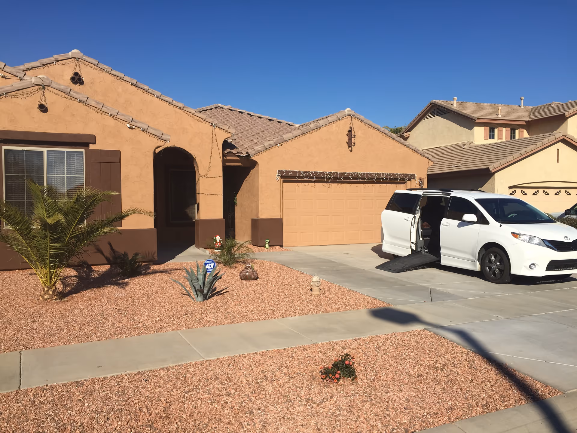 Front exterior view of a single-story house with a beige stucco finish and brown trim. The house has a tiled roof, a garage with a closed door, and a driveway where a white van with an open side door and a wheelchair ramp is parked. The front yard is landscaped with reddish gravel, small plants, and a palm tree. The sky is clear and blue.