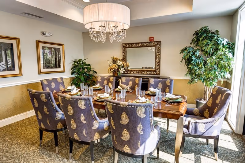 A well-lit dining room with a wooden table set for eight people. The chairs have a purple upholstery with a gold paisley pattern. The table is set with plates, napkins, glasses, and silverware. A large chandelier with crystal accents hangs above the table. There is a large mirror on the wall, two framed pictures, and two potted plants near the window letting in natural light.