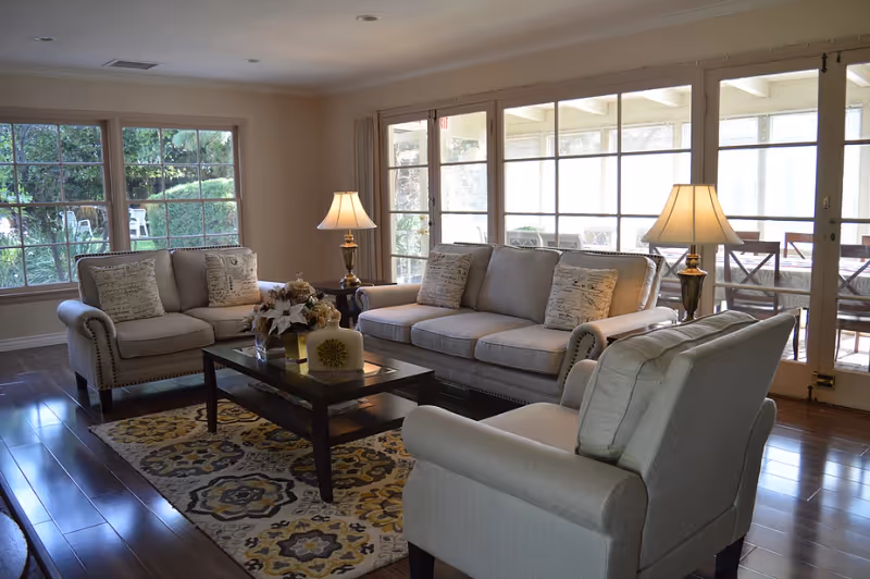 Sunlit living room with beige sofas and armchairs arranged around a coffee table on a patterned rug, flanked by lamps and large windows/doors to a patio.