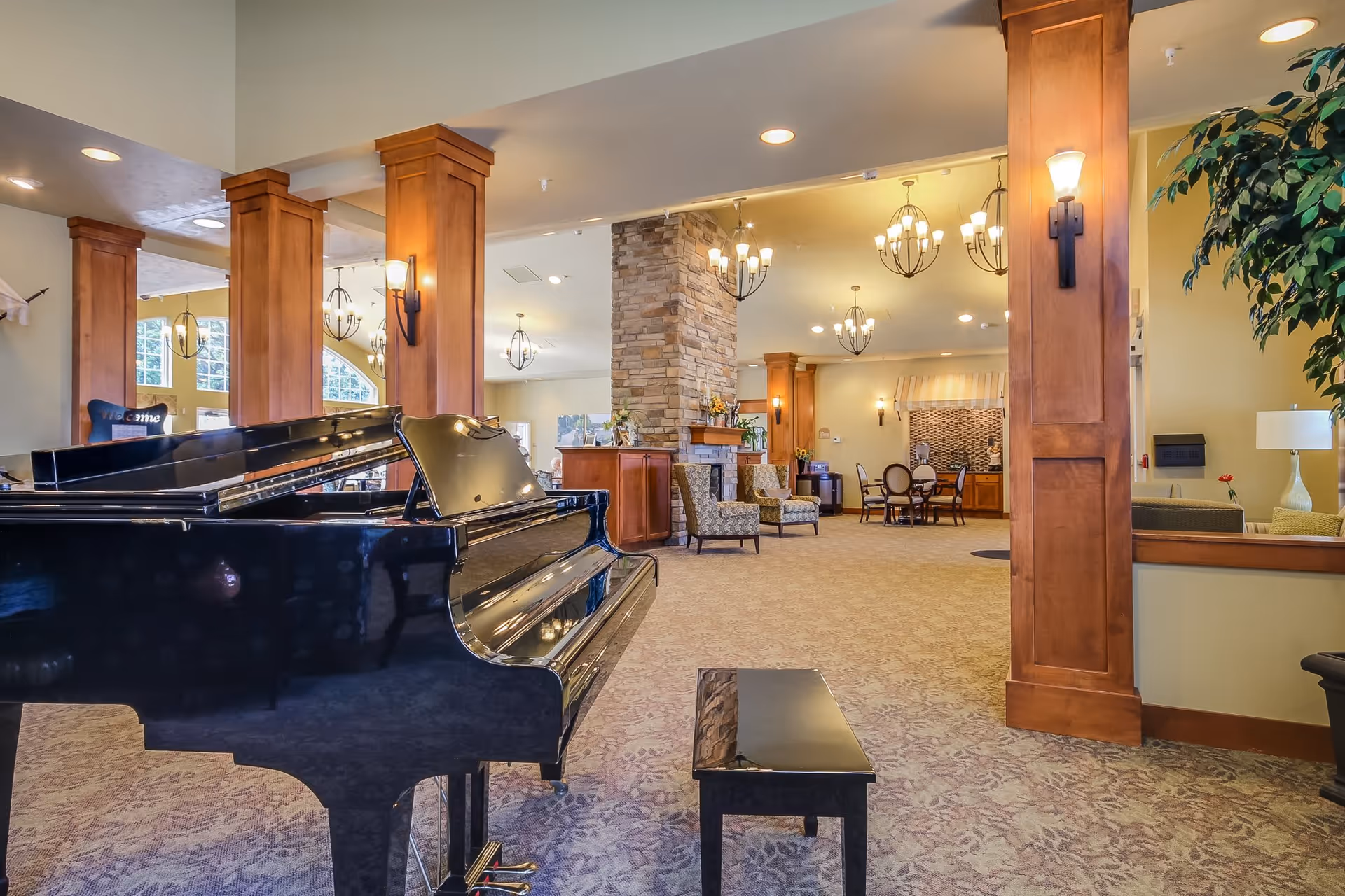 Spacious and well-lit common area in an assisted living facility featuring a black grand piano with a matching bench in the foreground, wooden columns with wall sconces, a stone fireplace surrounded by comfortable chairs, chandeliers hanging from the ceiling, and a dining area with tables and chairs in the background.